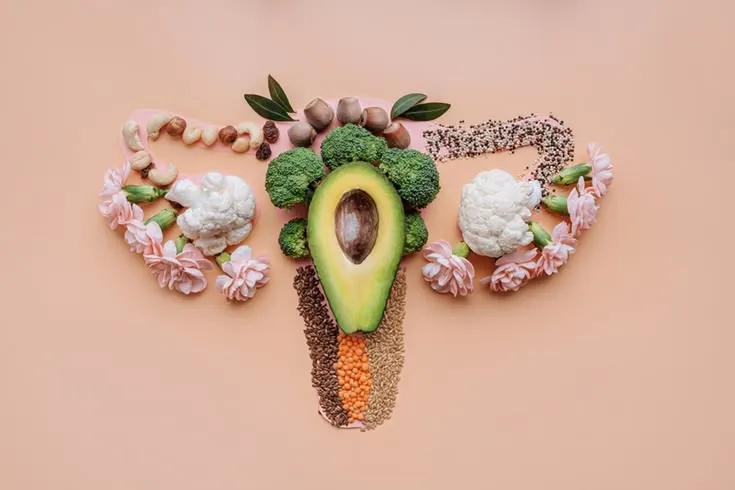 Apparato riproduttivo femminile composto da avocado, broccoli, cavolfiori, semi e frutta secca su fondo color pesca