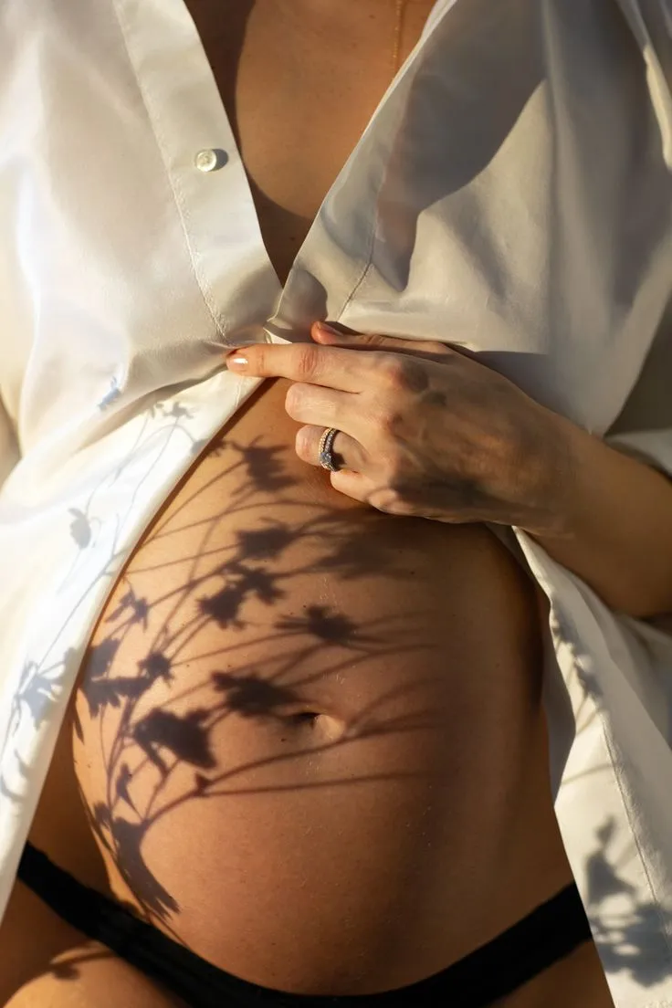 Pancione di una donna incinta in camicia bianca, con l’ombra di rami fioriti proiettata sulla pelle