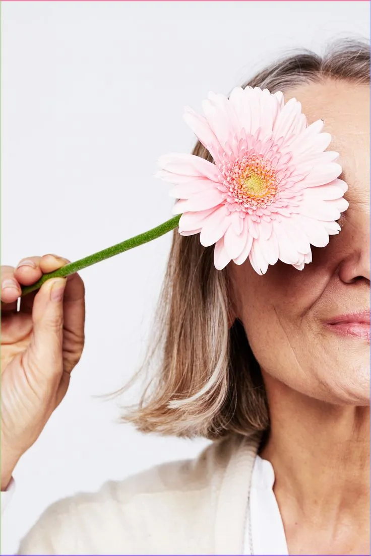 Donna con capelli grigi che si copre un occhio con una gerbera rosa